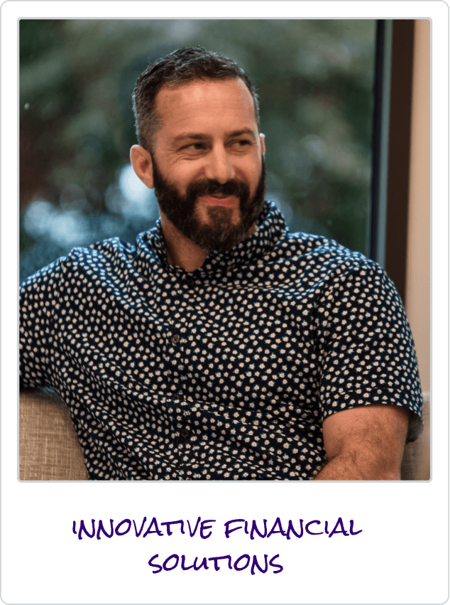 Man with beard wearing a patterned shirt sitting on a couch, smiling and looking to the side.