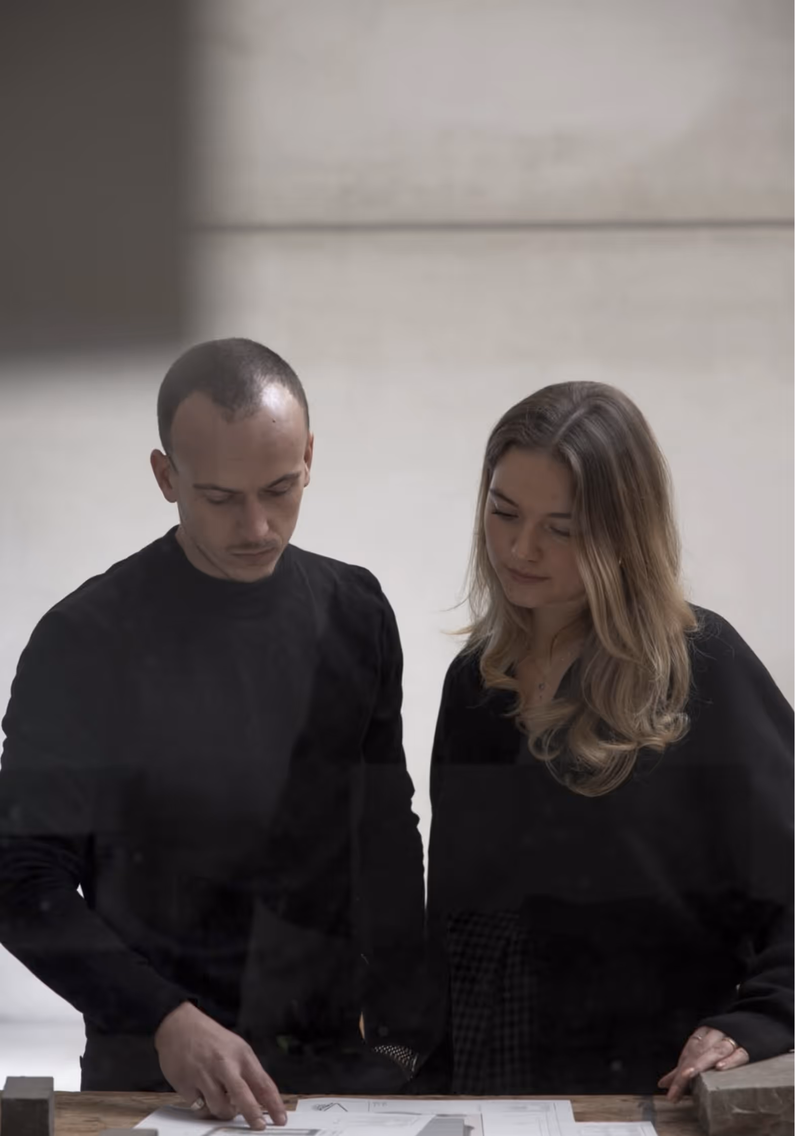 Twee mensen (Olivier en Gaëlle) bekijken plannen of tekeningen op een tafel in een rustige, neutrale ruimte.
