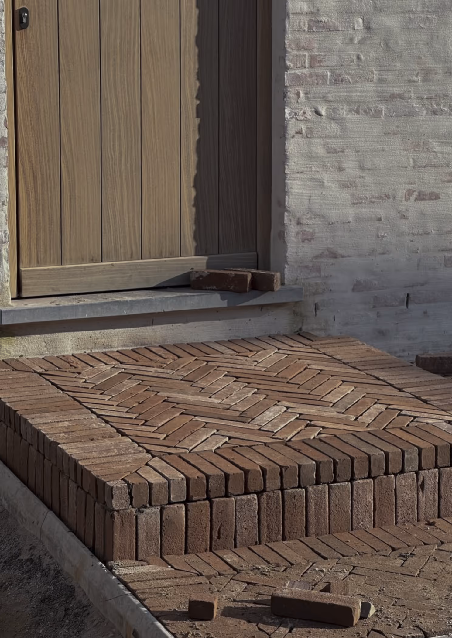 Voordeur van hout met een verhoogd terras van bakstenen in visgraatmotief en enkele losse bakstenen erop.