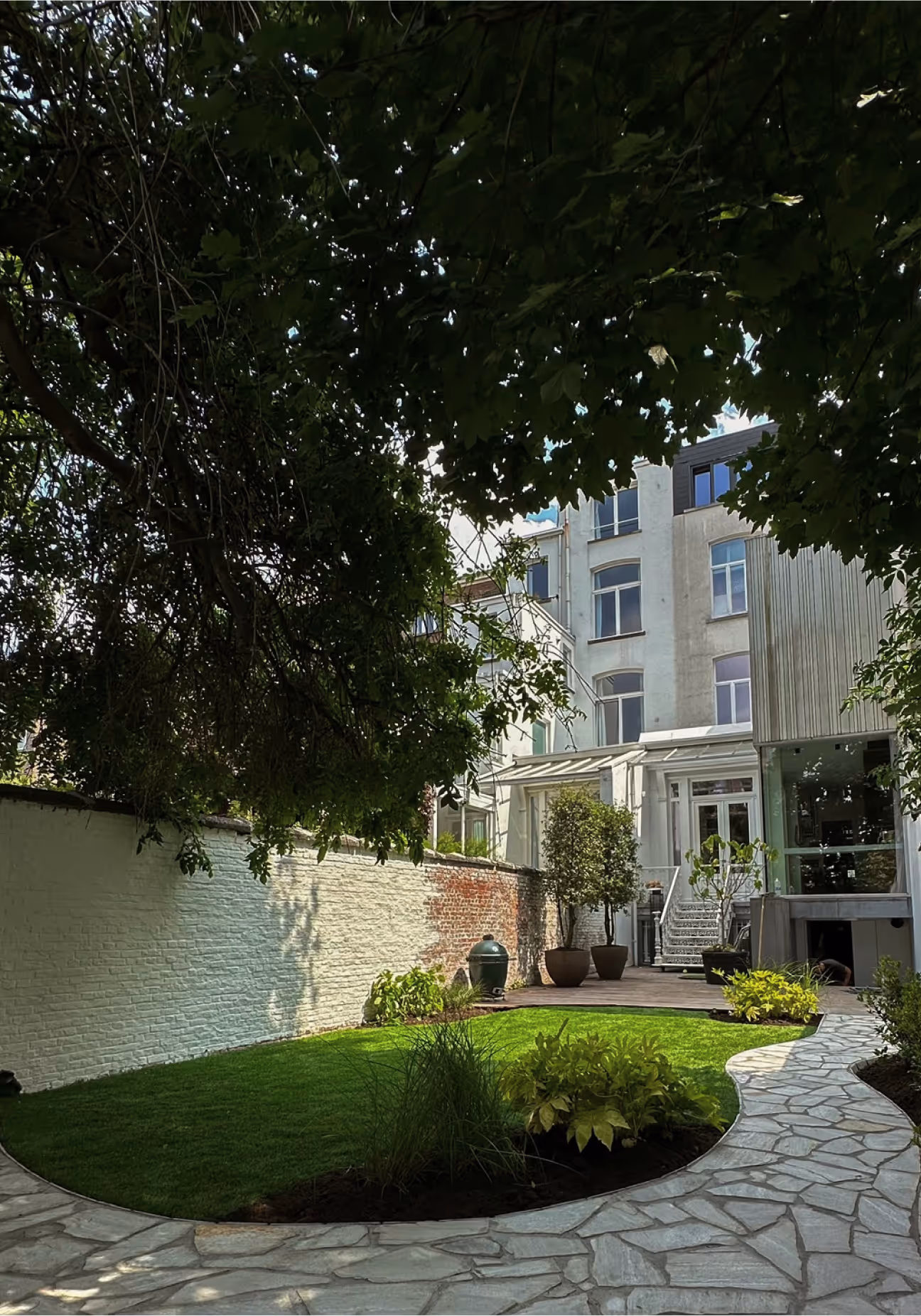 Tuin met een rond grasveld, beplanting langs het pad, een wit geschilderde bakstenen muur en een woonhuis met glazen veranda op de achtergrond.