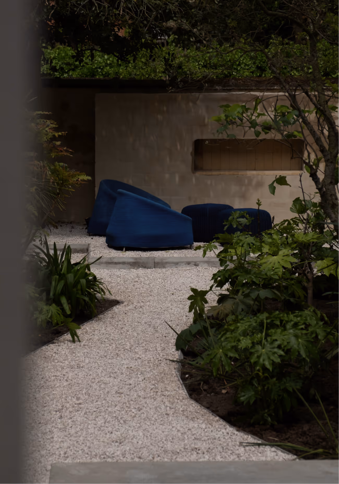Tuinpad van grind leidt naar een buitenzitgedeelte met twee blauwe stoelen en een bijpassende poef, omringd door groene planten.