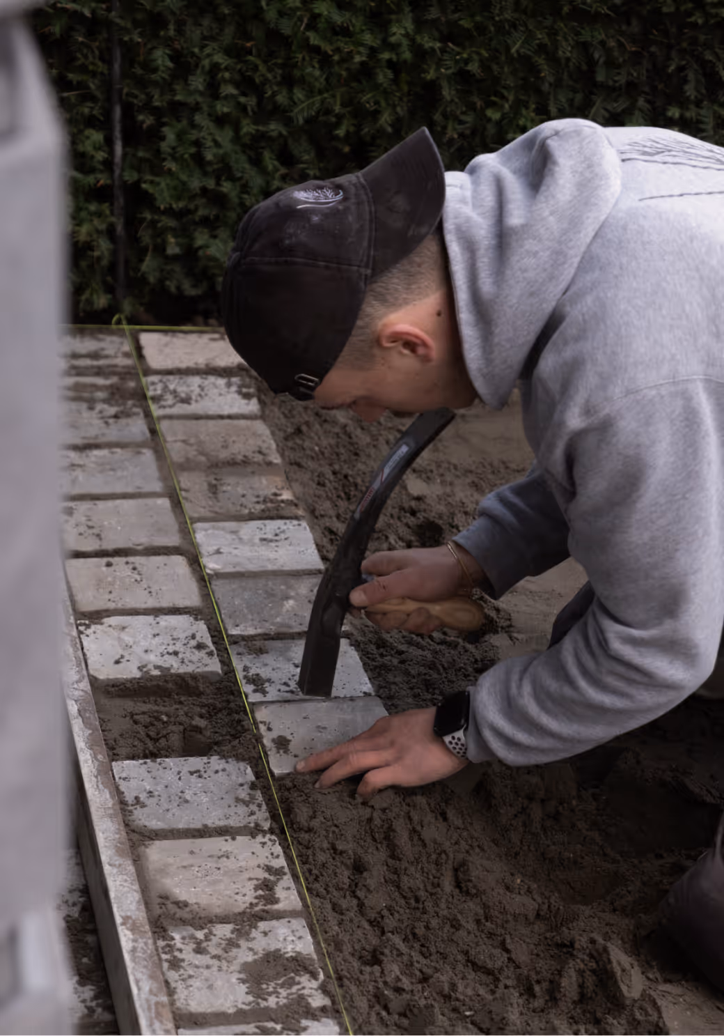 Man (Olivier) met zwarte pet en grijze hoodie legt bestratingstenen met troffel en touwlijn op een zandbed.