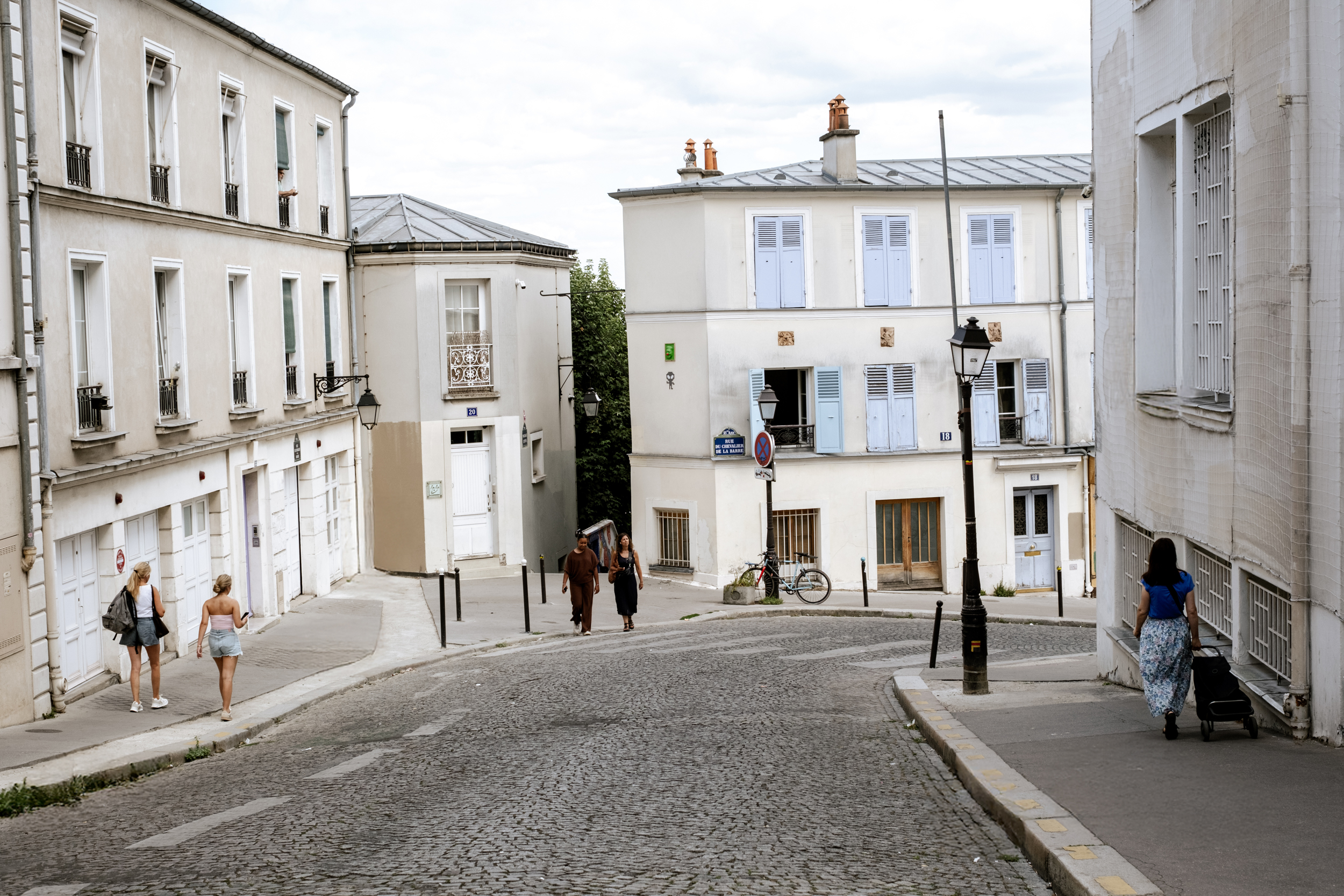 Rue pavée en pente avec des piétons marchant près de bâtiments résidentiels à fenêtres bleues.