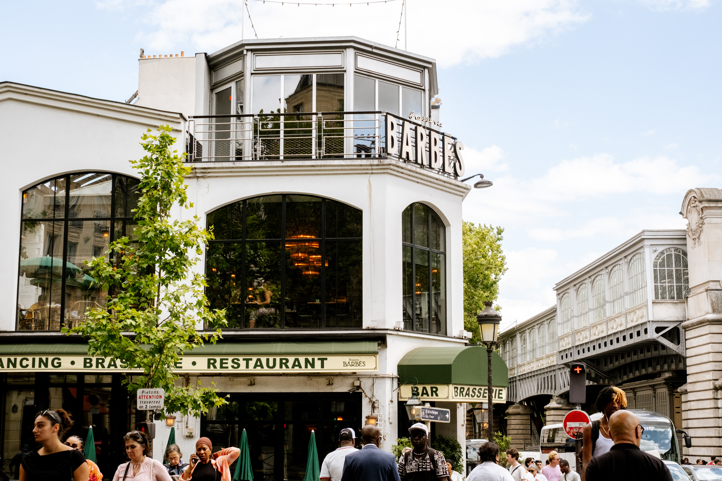 Façade de la brasserie Barbès avec grandes fenêtres, arbres, lampadaire et foule de passants.