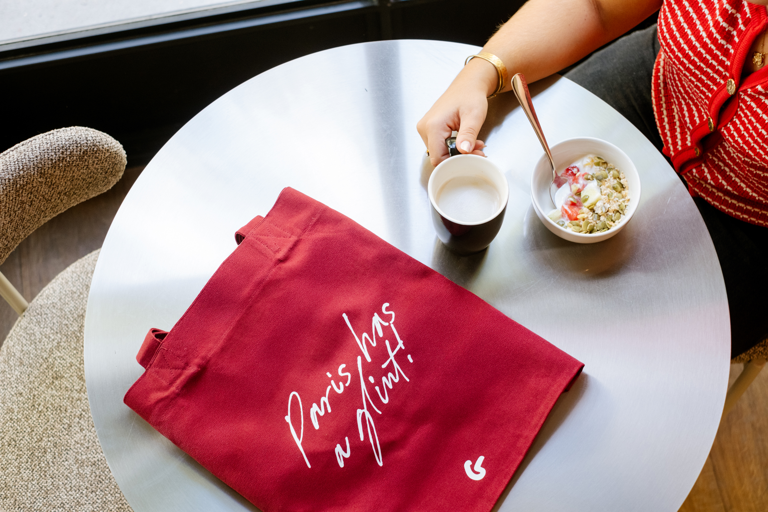 Sur une table ronde, un sac rouge, une main tenant une tasse à café et un bol de yaourt avec fruits et graines.