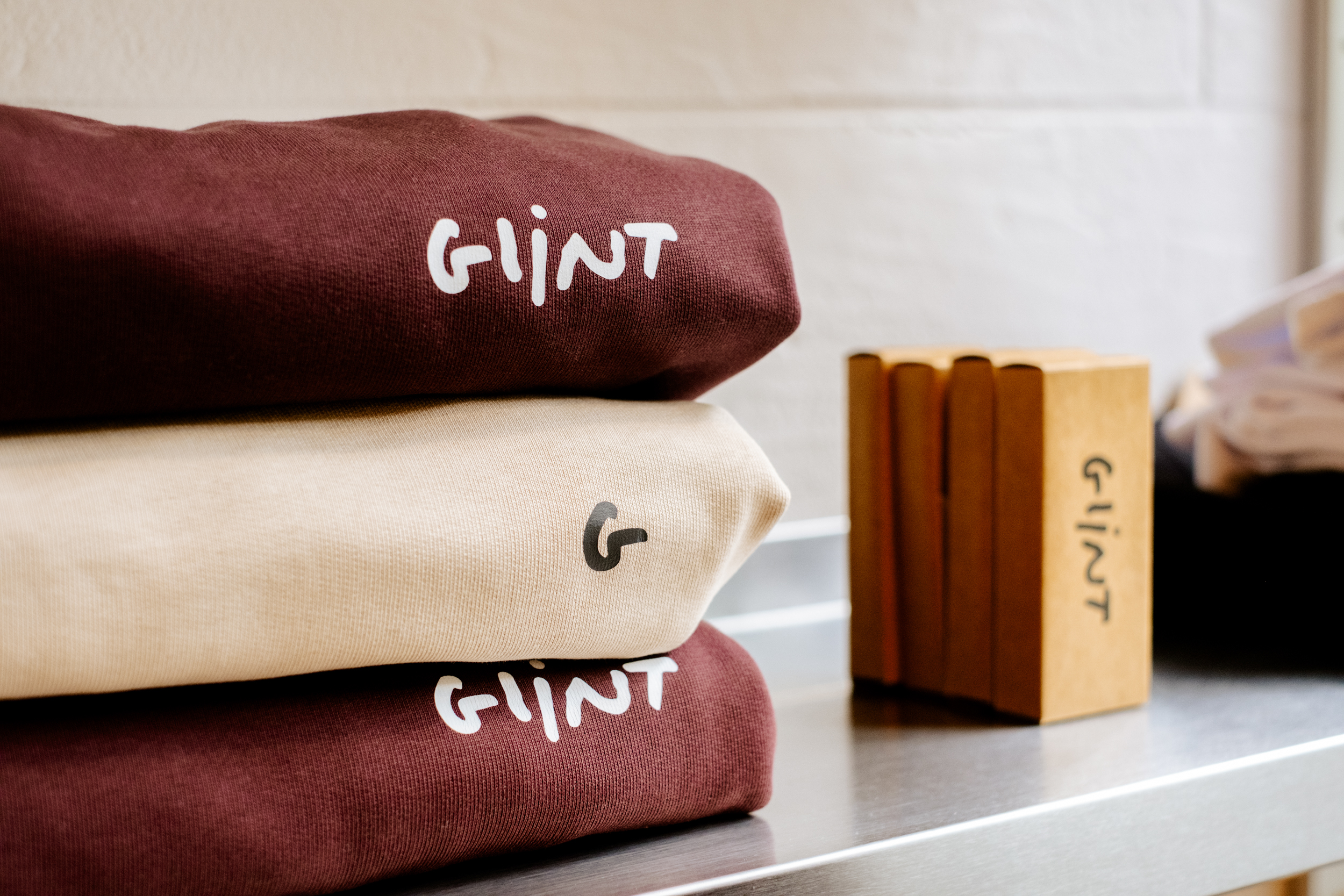 Pile de trois sweat-shirts pliés avec le logo Glint, à côté de boîtes en carton Glint sur une étagère métallique.