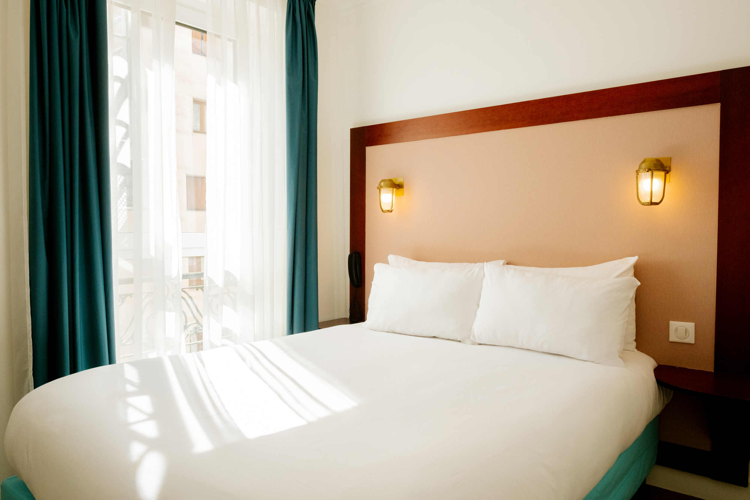 Chambre lumineuse avec un grand lit double blanc, deux lampes murales allumées et rideaux bleus.