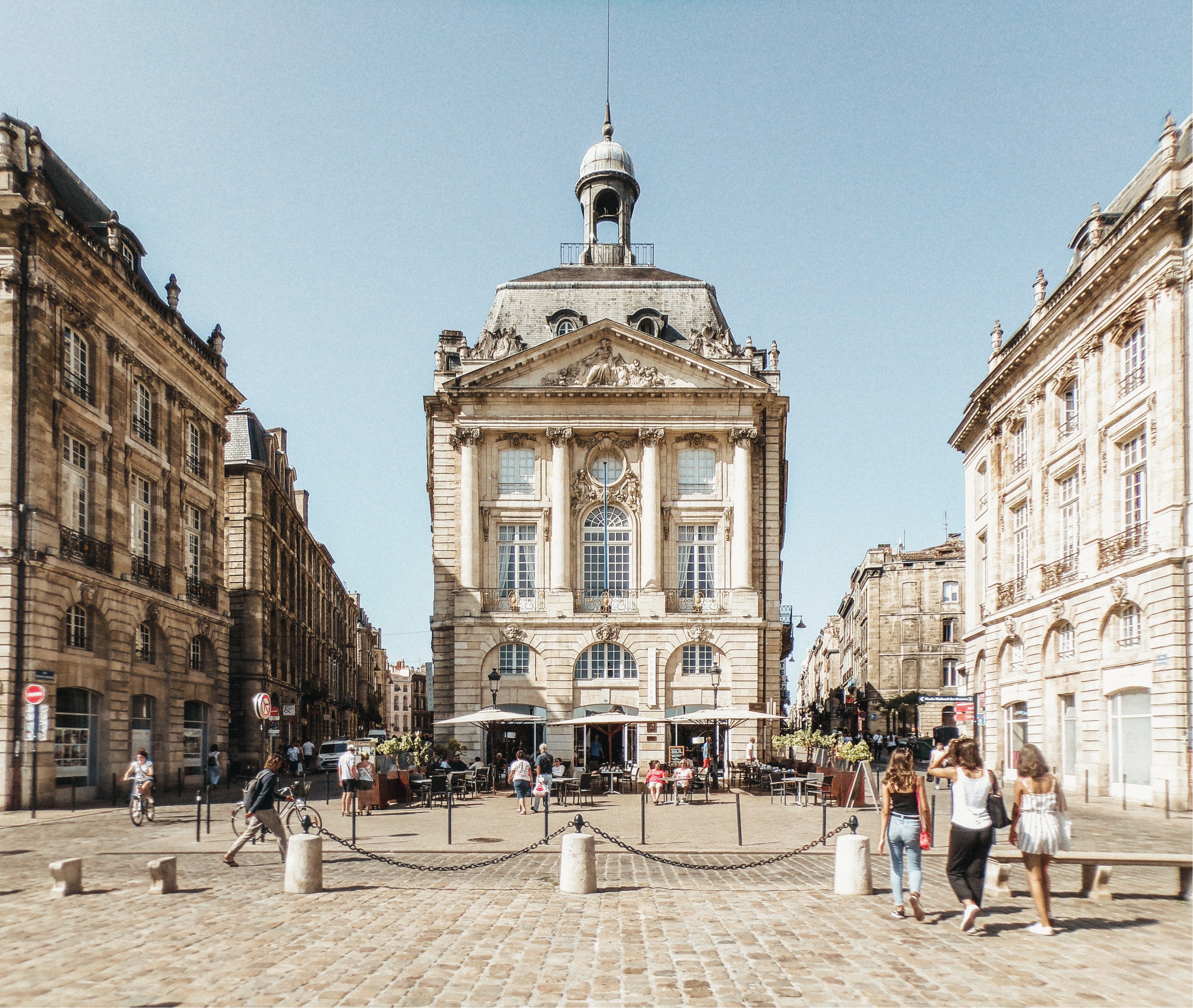 Place avec bâtiment ancien, terrasse de café et personnes marchant sur pavé sous ciel bleu.