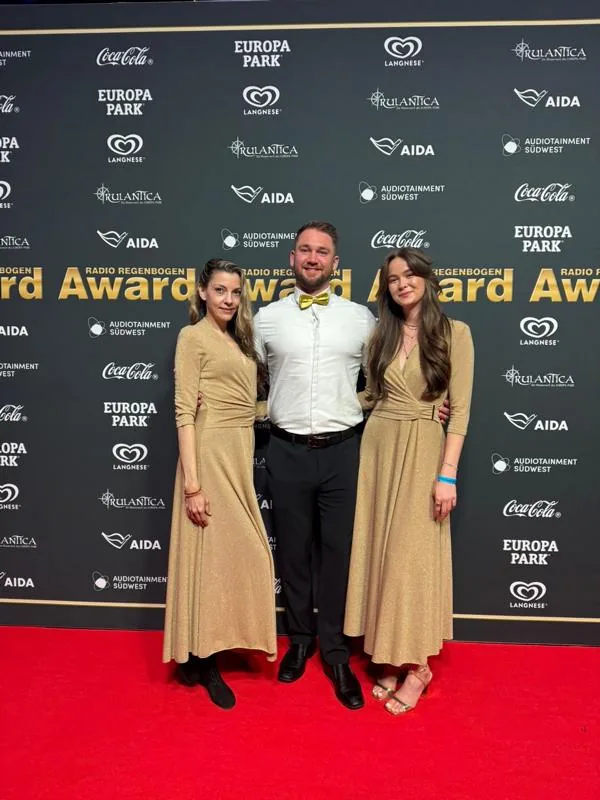 Zwei Promoterinnen in goldfarbenen Kleidern und ein Kollege mit gelber Fliege auf dem roten Teppich vor der Award-Wand des Radio Regenbogen Award im Europa-Park Rust