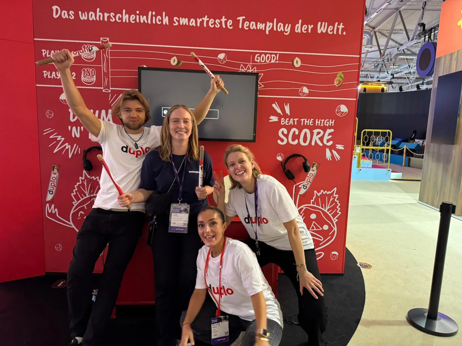 Vier Personen mit Trommelstöcken vor der Drum-’n’-Beat-Wand am duplo-Stand auf der gamescom 2026 in Köln