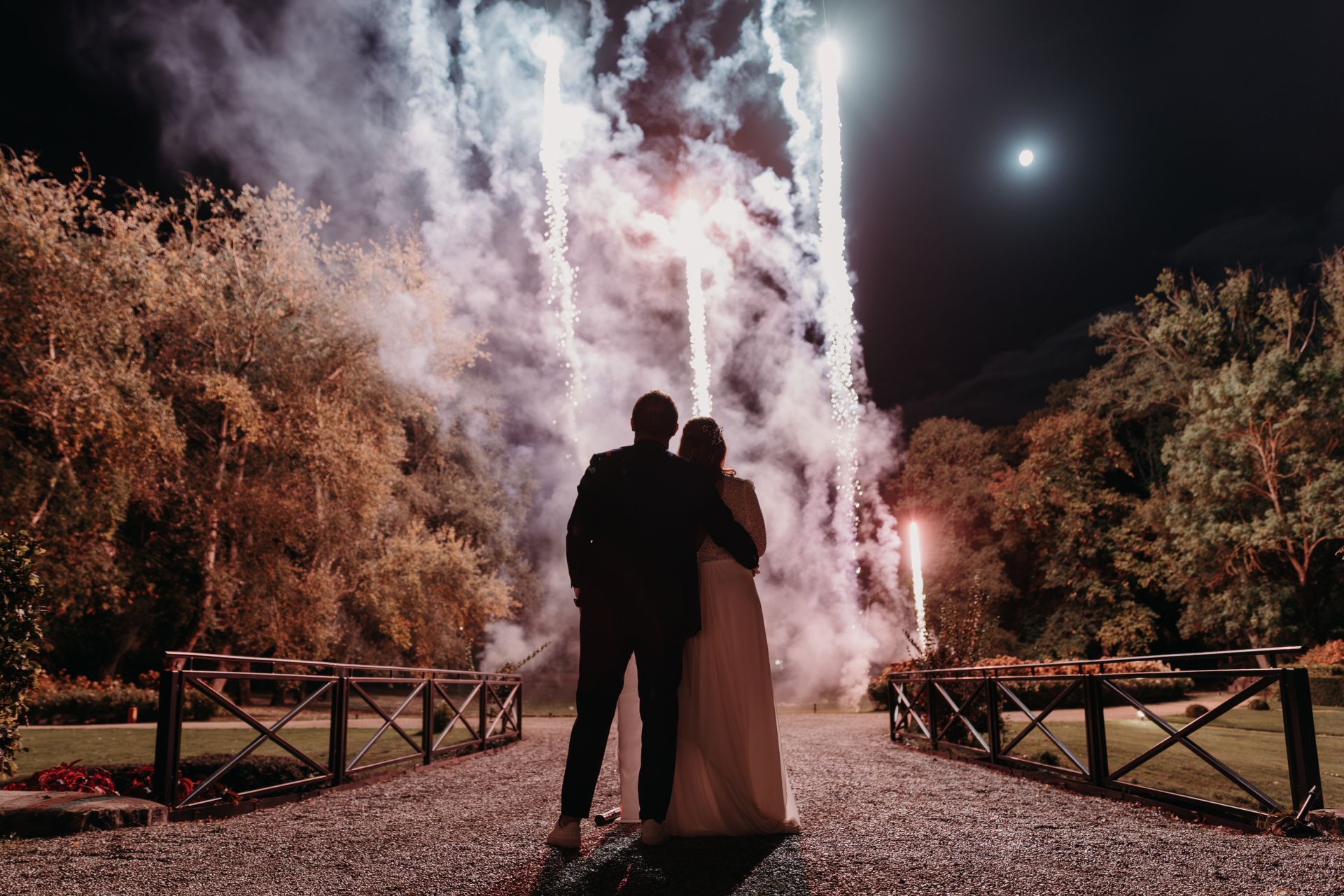 Couple de mariés enlacés de dos admirant des chandelles pyrotechniques blanches s'élevant dans le ciel nocturne au-dessus d'un parc arboré, sous la lumière de la lune, spectacle feux d'artifice mariage sur mesure réalisé par Wagnon