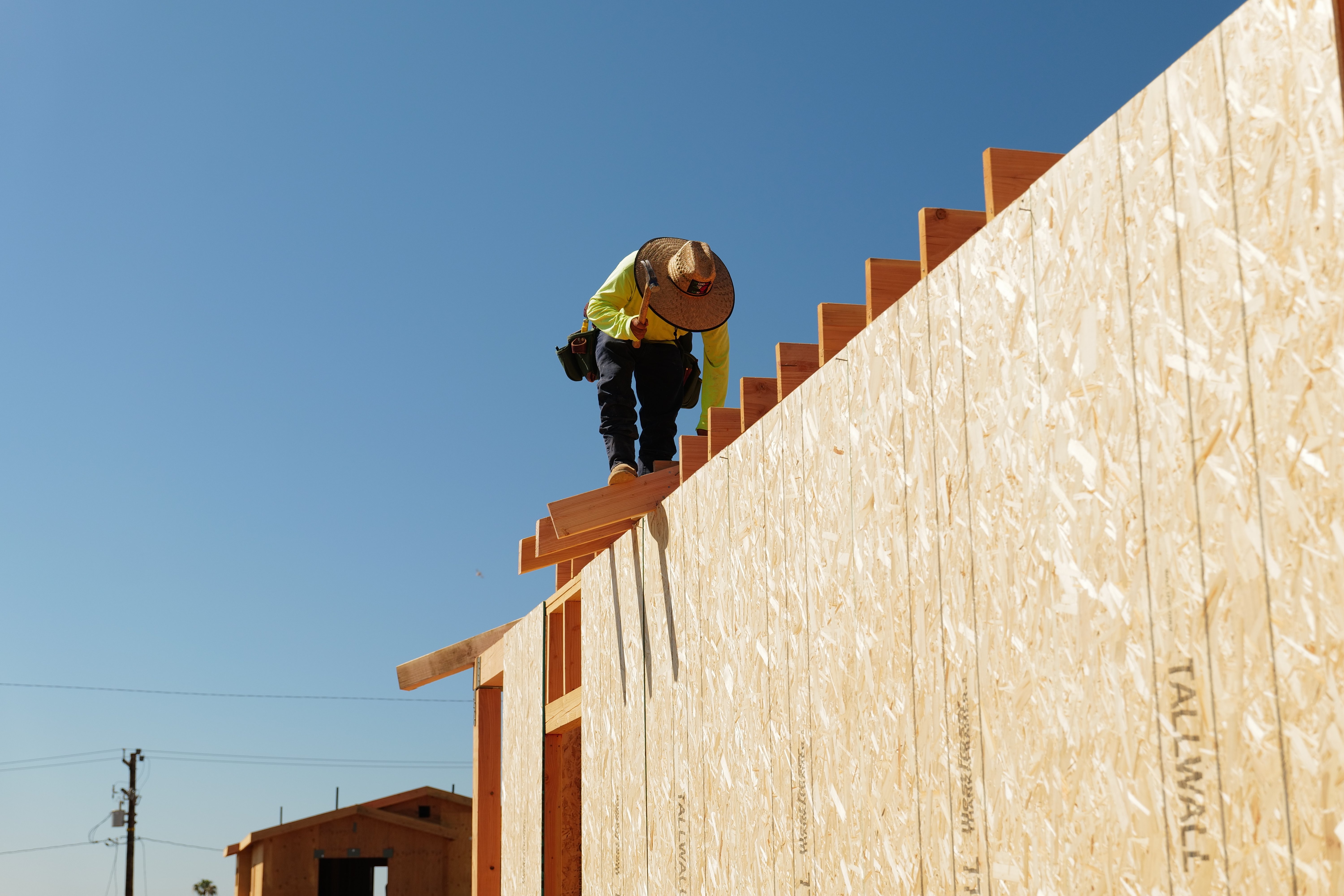A worker on the top edge of tall wall sheathing on an Altadena rebuild, with an overhead power line and wood utility pole behind under a clear sky