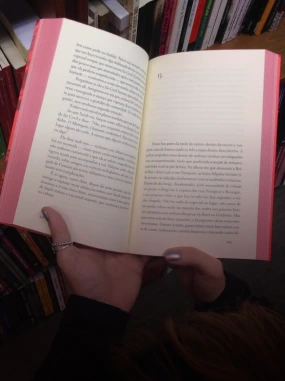 Pessoa segurando um livro aberto com texto impresso em português, em uma biblioteca com estantes e livros ao fundo.