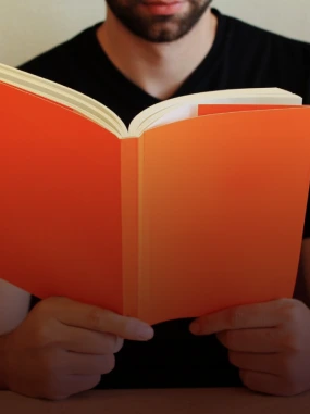 Pessoa lendo um livro de capa laranja, segurando-o com ambas as mãos.