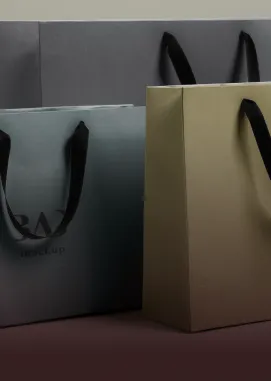Três sacolas de papel com alças pretas posicionadas lado a lado, uma cinza, uma dourada e outra cinza escura ao fundo.