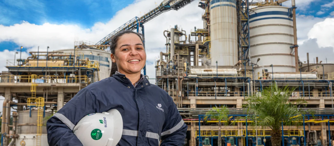 Mulher vestindo uniforme azul da �ڰ������� sorrindo, segurando capacete branco em frente a uma instalação industrial com silos e tubulações.