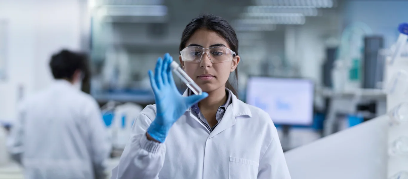 Pesquisadora em jaleco branco e óculos de proteção segurando um tubo de ensaio em laboratório moderno.