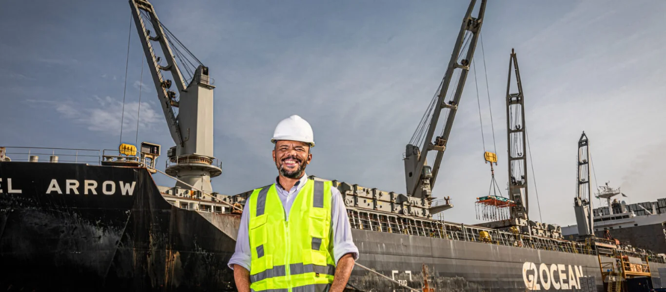 Homem com capacete e colete de segurança amarelo sorrindo em frente a um grande navio carga com guindastes operando.