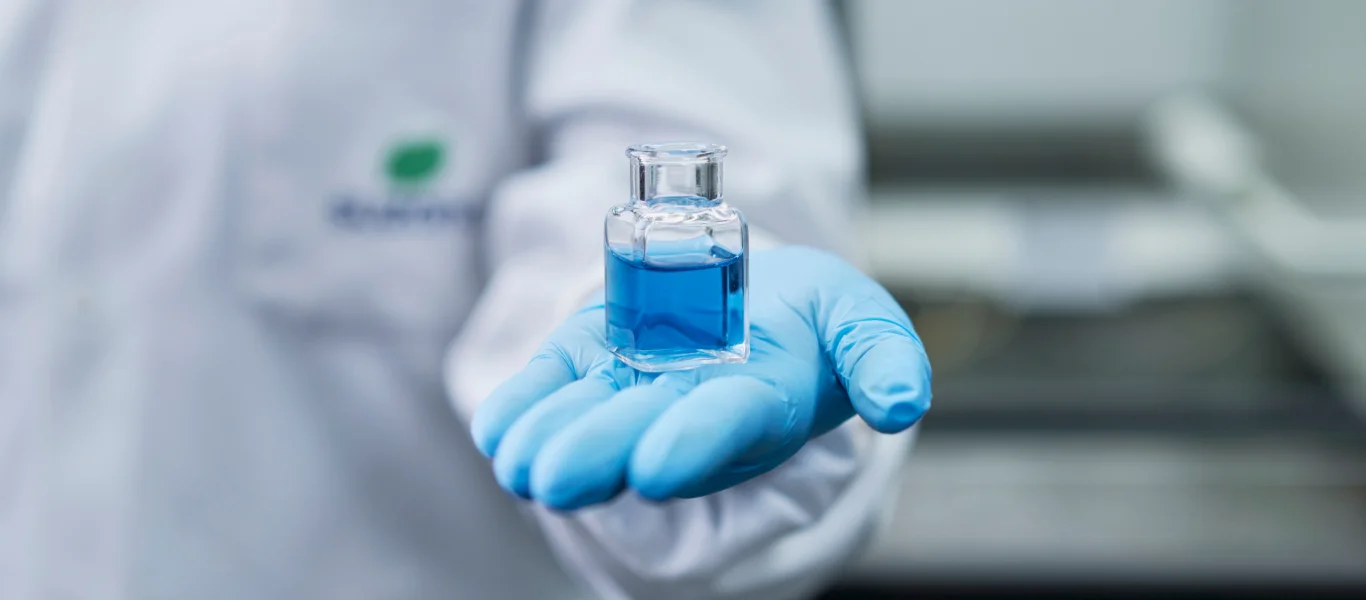 Mão com luva azul segurando um frasco pequeno com líquido azul em ambiente laboratorial.