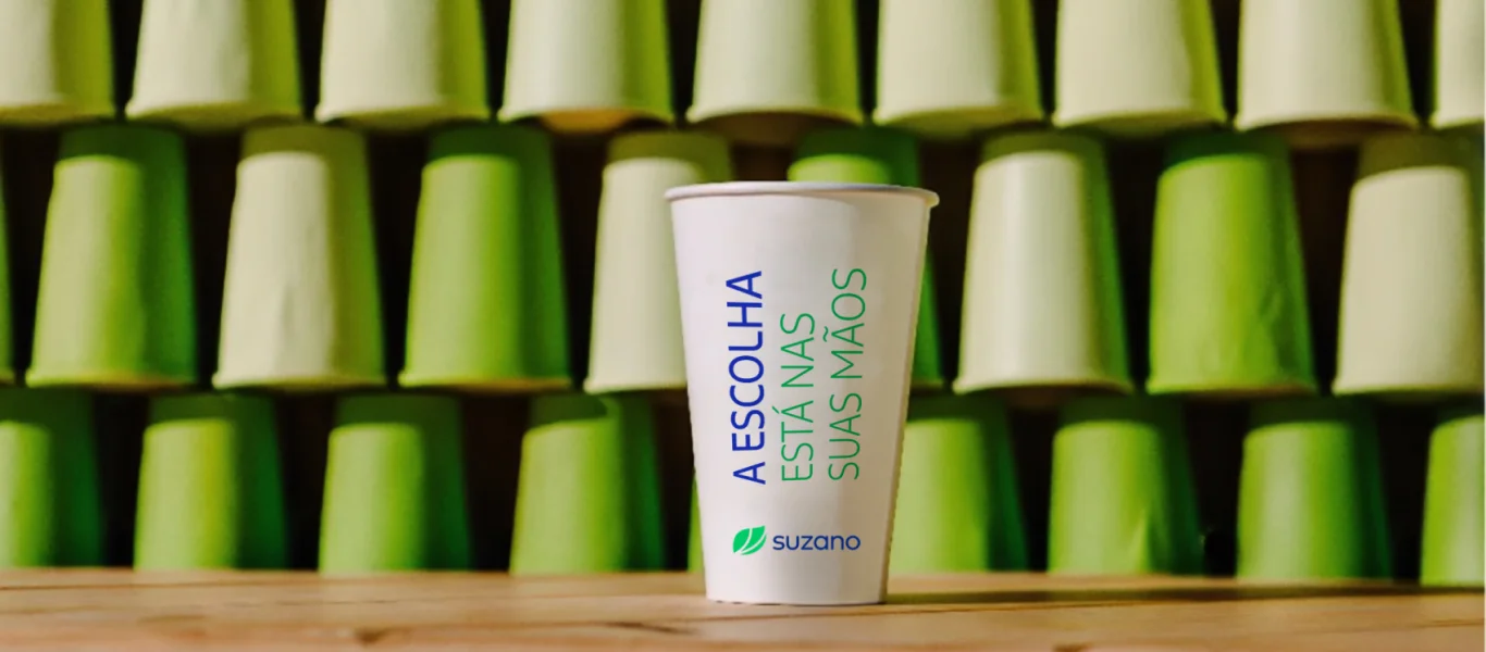 Copo branco de papel com a frase 'A escolha está nas suas mãos' e o logo da Suzano, com fundo de copos verdes empilhados.