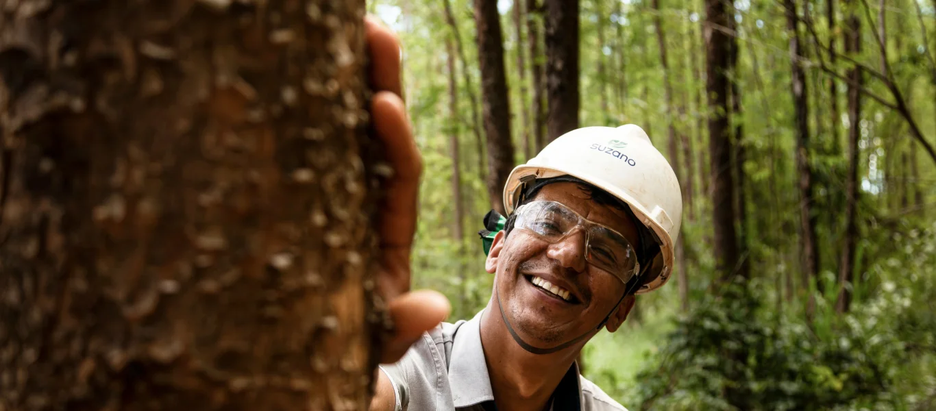 Homem sorridente usando capacete e óculos de proteção encostado em uma árvore na floresta.