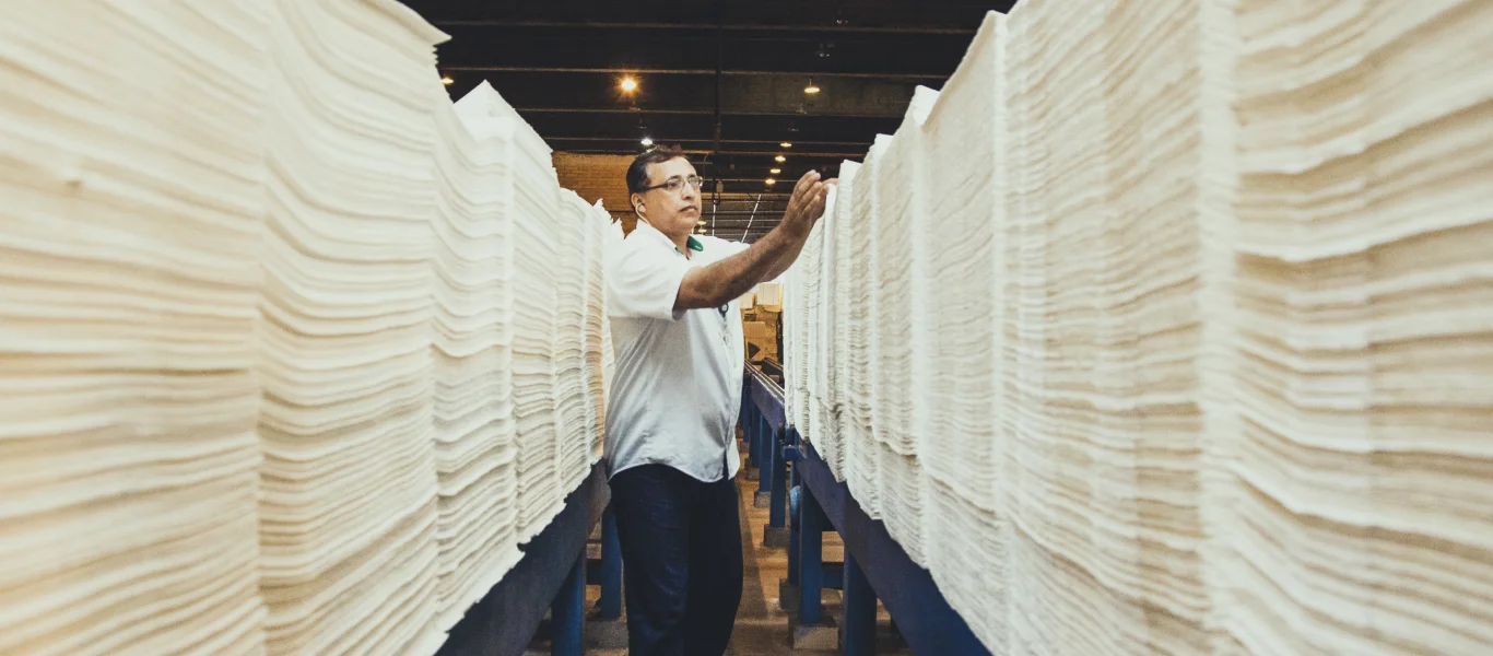 Homem inspecionando grandes pilhas de papel branco empilhado em uma fábrica.