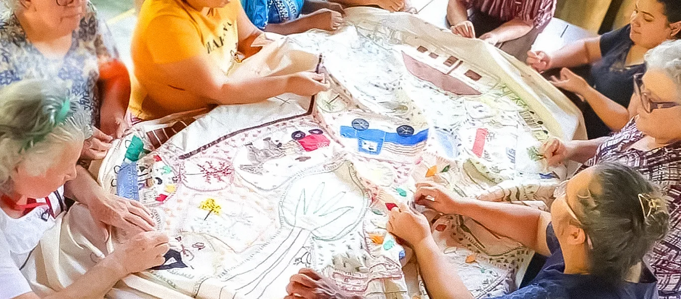 Grupo de mulheres sentadas ao redor de uma mesa bordando um grande tecido com desenhos coloridos.