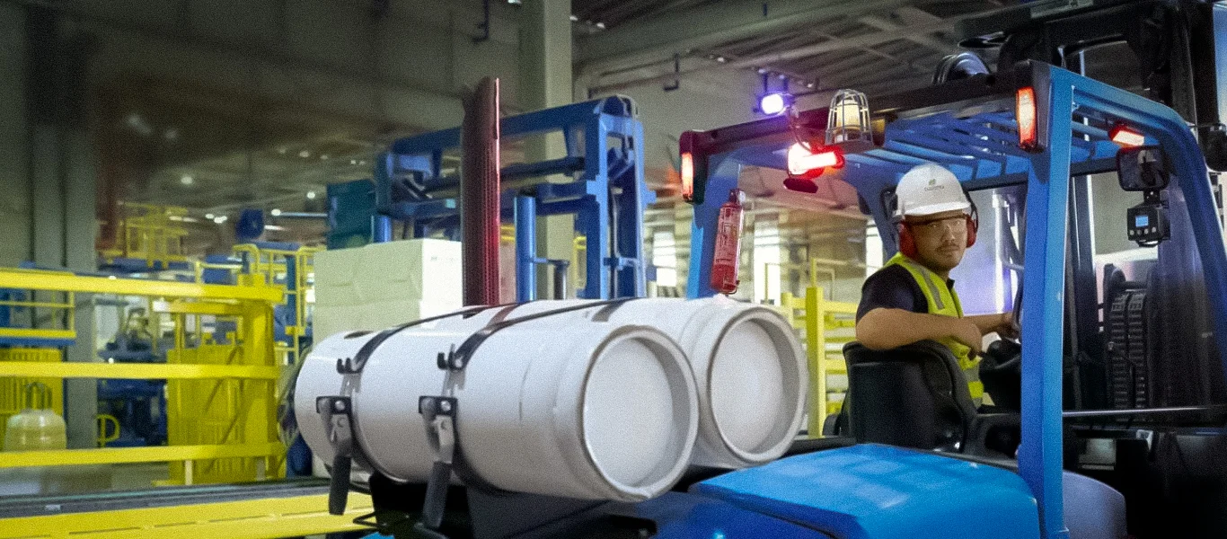 Operador com capacete e colete de segurança dirigindo empilhadeira azul dentro de um ambiente industrial com barris brancos.