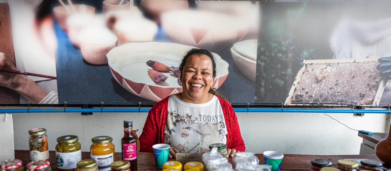 Mulher sorridente sentada atrás de uma mesa com diversos potes de conservas e frascos, com um banner colorido e desfocado ao fundo.