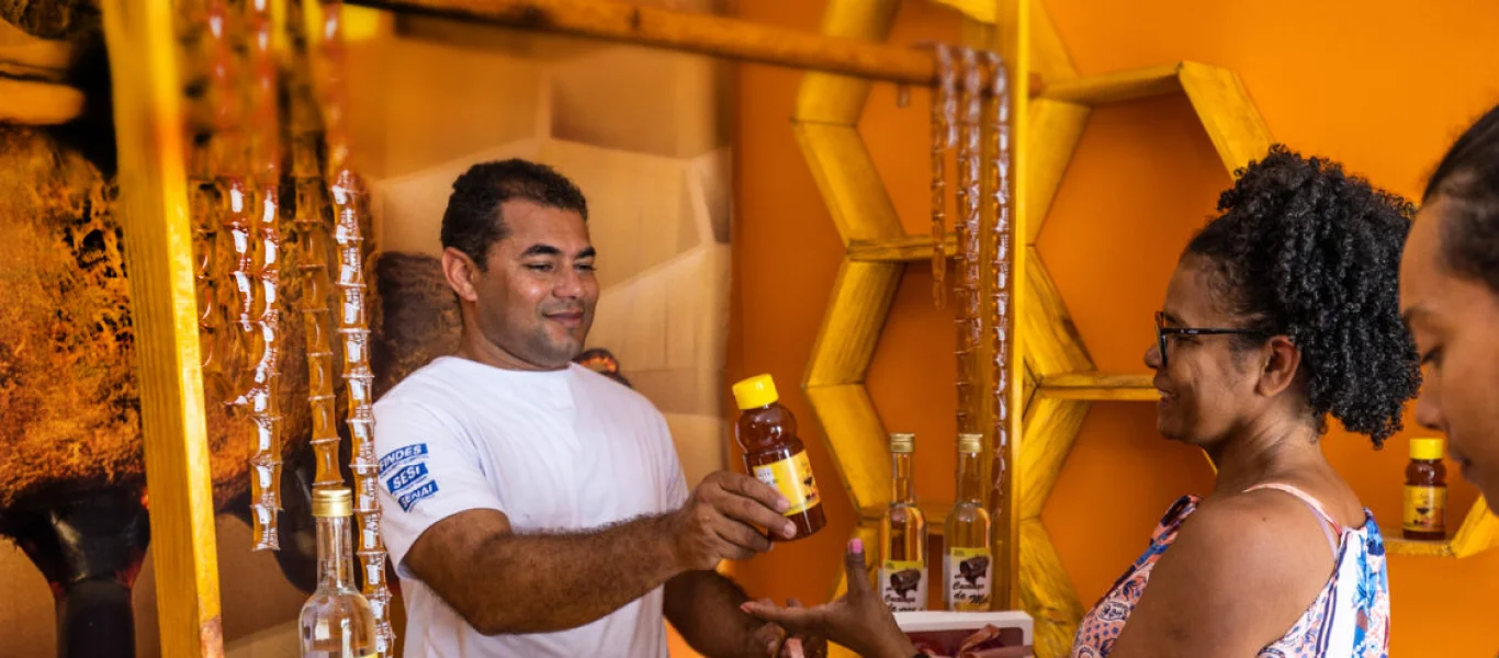 Homem entregando uma garrafa a uma mulher em um ambiente decorado com elementos que remetem a colmeias e produtos em frascos.