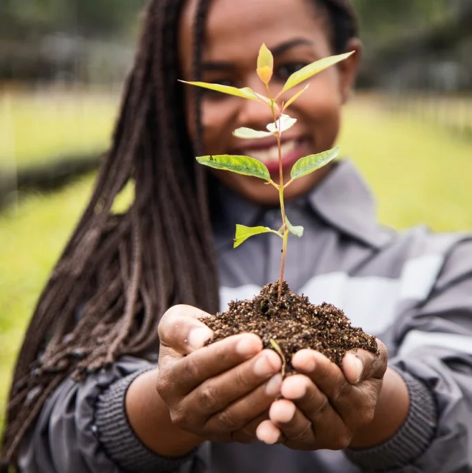 Pessoa sorridente segurando um pequeno broto de planta com terra nas mãos estendidas.