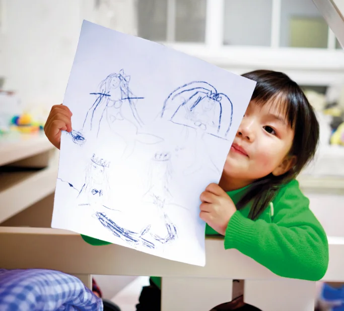 Criança segurando e mostrando um desenho de sereias feitas à mão em papel branco.