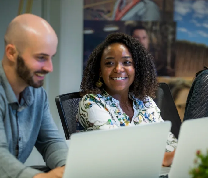Duas pessoas sorrindo enquanto trabalham em laptops em uma mesa de escritório.