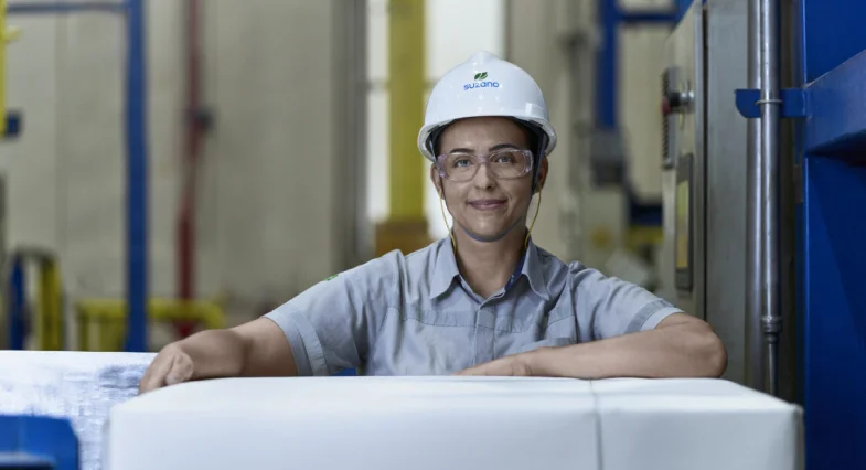 Mulher com capacete branco e óculos de proteção em fábrica, sorrindo e apoiada em blocos empilhados.