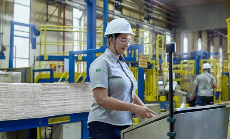 Mulher usando capacete e óculos de segurança operando máquina em ambiente industrial com equipamentos e prensas ao fundo.