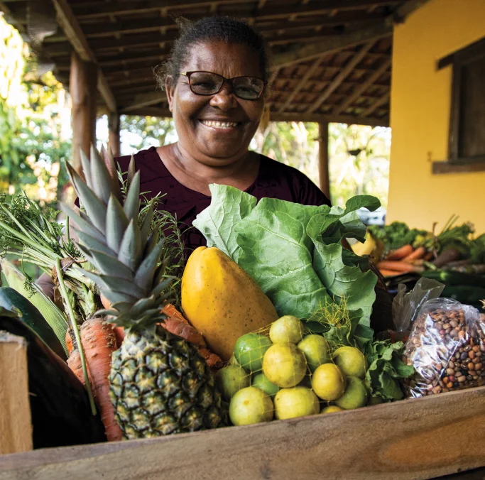 Mulher sorridente segurando uma caixa de madeira cheia de frutas e legumes frescos, como abacaxi, mamão, alface, cenoura e limões.