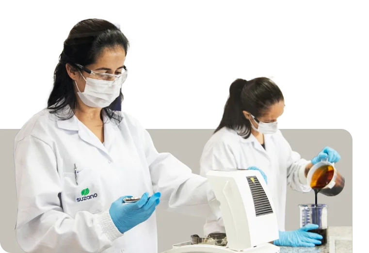Duas mulheres cientistas em jalecos brancos, máscaras e luvas azuis, uma segurando um frasco e a outra despejando líquido marrom numa proveta em laboratório.