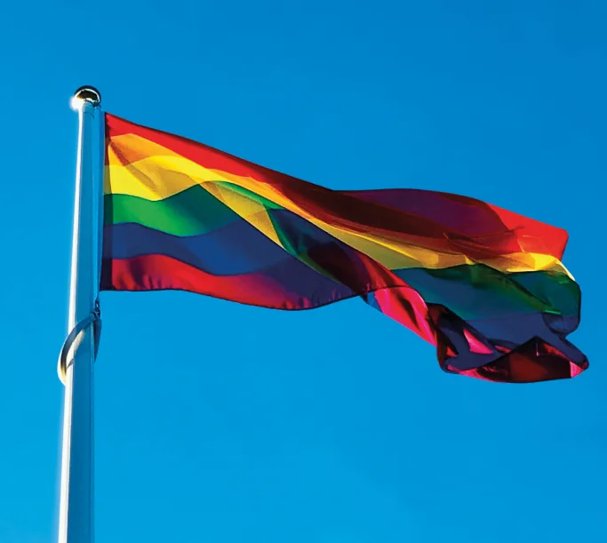 Bandeira do arco-íris tremulando ao vento contra um céu azul claro.