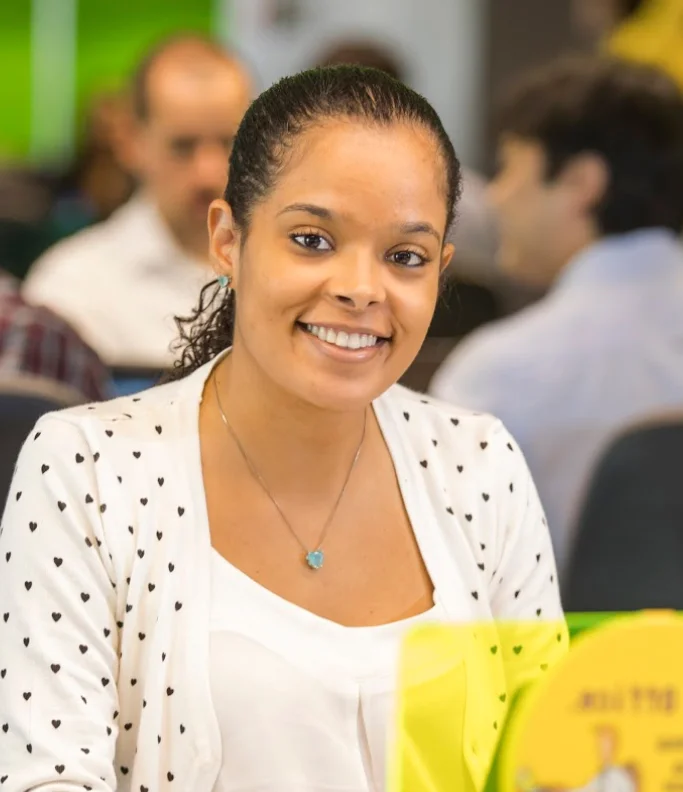 Mulher sorridente com suéter branco de corações pretos sentada em ambiente de escritório com pessoas ao fundo.