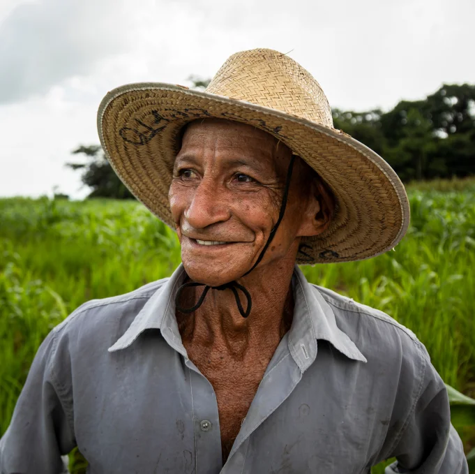 Homem sorridente usando chapéu de palha em campo verde ao ar livre.