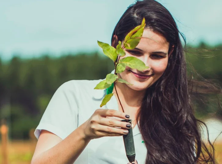 Mulher sorridente segurando uma muda de planta ao ar livre em um ambiente natural.