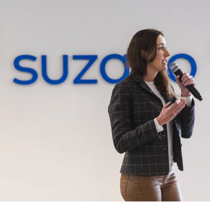 Mulher falando ao microfone em frente a uma parede com o logotipo azul da empresa '�ڰ�������'.