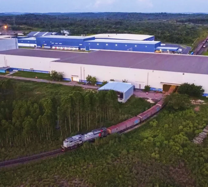 Trem de carga passando por trilhos perto de grandes galpões industriais cercados por áreas verdes.