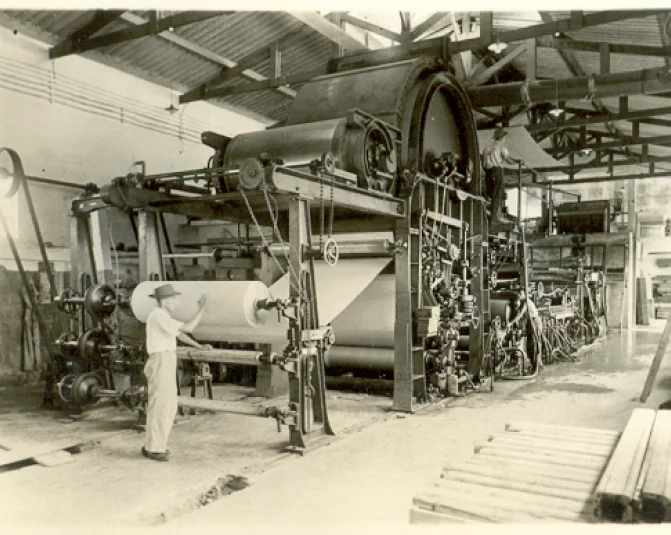 Homem operando uma grande máquina industrial antiga para produção de papel em uma fábrica.