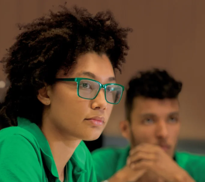Mulher com óculos verdes e cabelo cacheado, vestindo camisa verde, em um ambiente interno, com homem desfocado ao fundo.