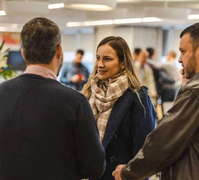 Mulher sorrindo conversa com dois homens em um ambiente interno com várias pessoas ao fundo.