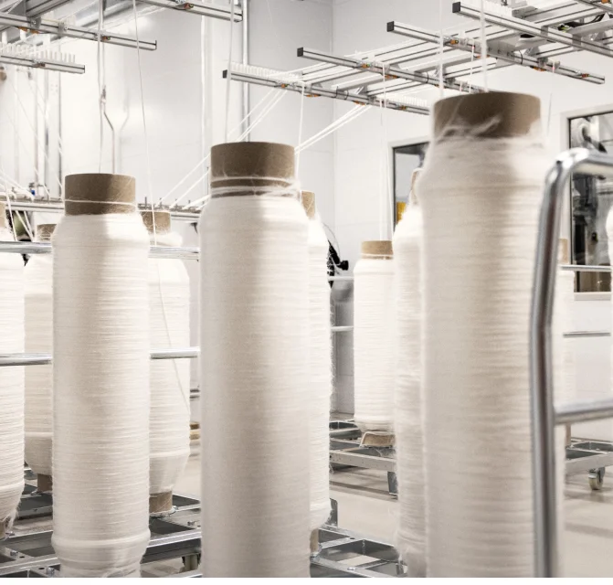 Múltiplos carretéis altos de fios brancos em uma fábrica têxtil com estrutura metálica e iluminação branca.