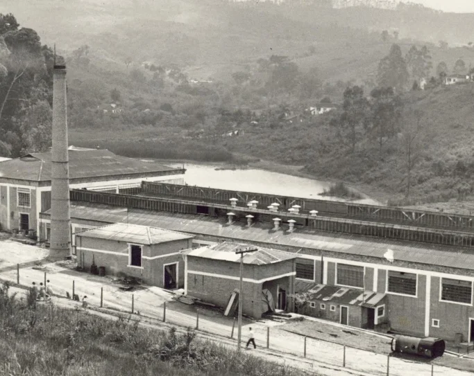 Foto em preto e branco de uma fábrica com chaminé alta, vários prédios industriais e uma estrada ao lado, cercada por área florestal e colinas ao fundo.