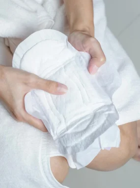 Mãos segurando um absorvente menstrual branco sobre uma perna coberta por toalha branca.