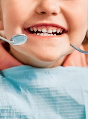 Criança sorrindo durante exame odontológico com espelho e sonda de dentista próximos à boca.